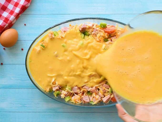 Pouring eggs over hash browns, ham, cheese, and vegetables for egg casserole