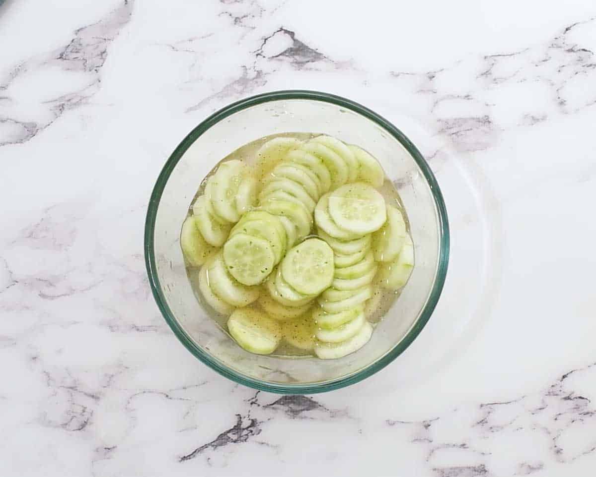 sliced cucumbers being marinated in vinegar in a glass bowl
