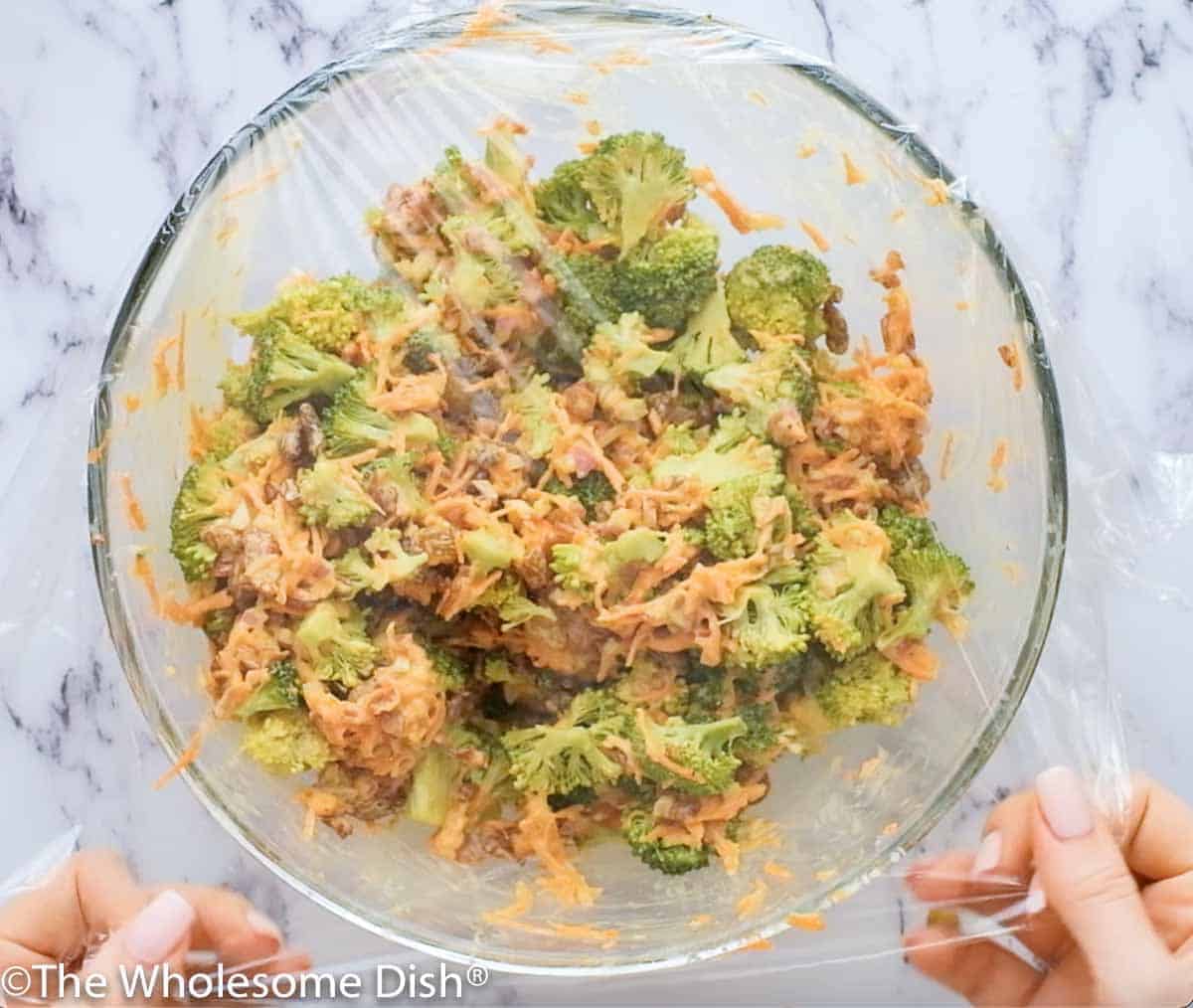 plastic wrap being placed over a large bowl of broccoli salad