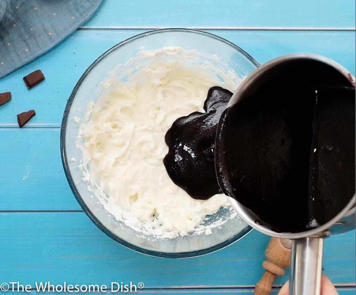 melted butter and chocolate being poured into a mixing bowl with cream cheese and powdered sugar