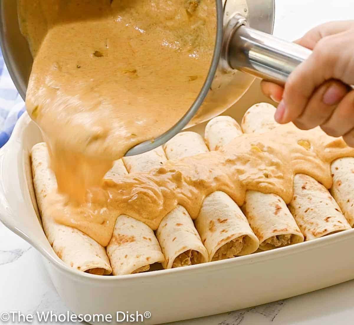 Baking dish full of rolled up chicken enchilada with sour cream sauce being poured over it.