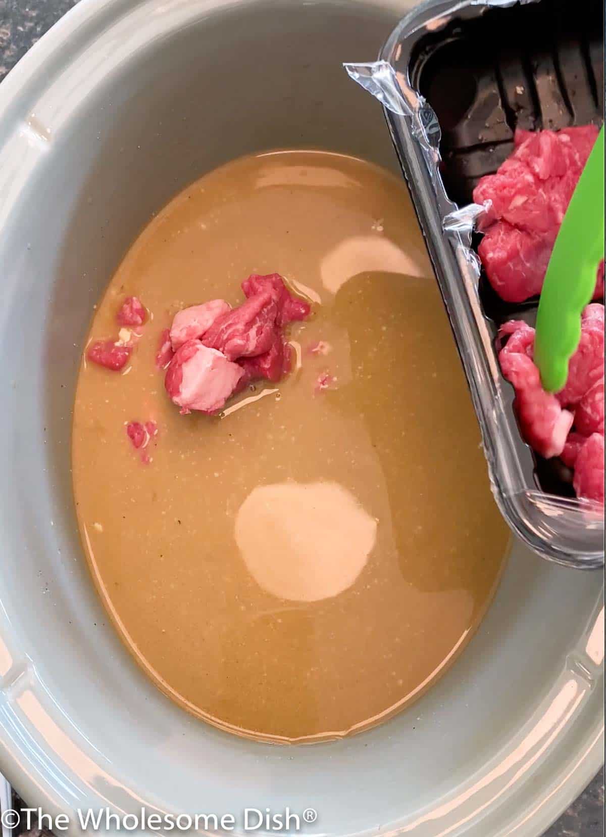 Uncooked beef stew meat being added to broth in a slow cooker.