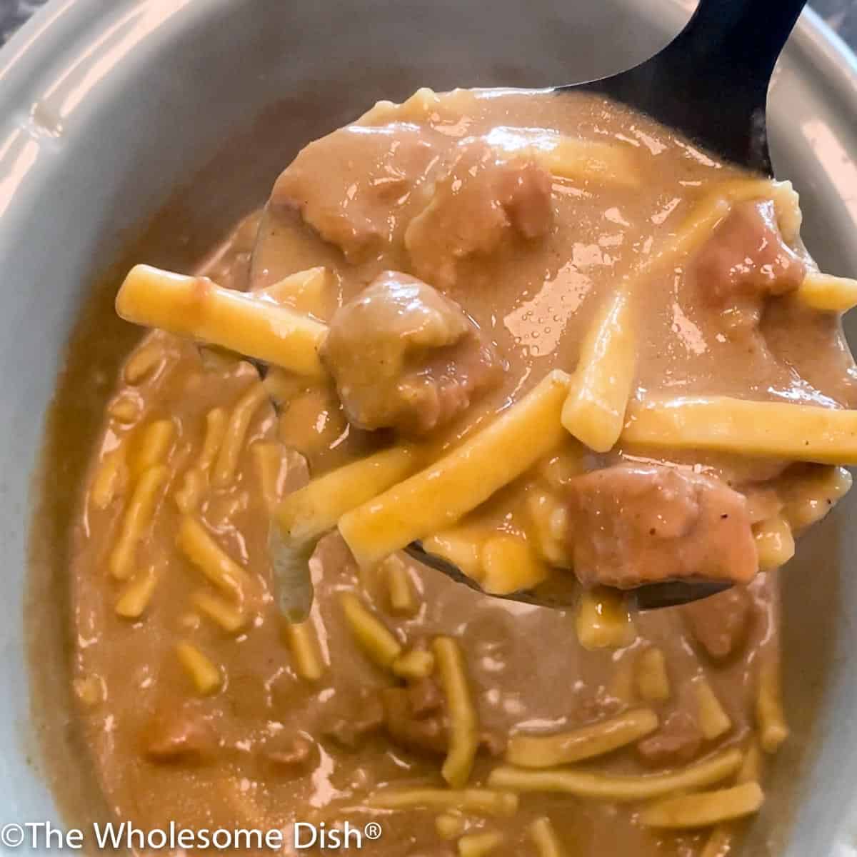 A ladle full of beef & noodles being lifted out of a crock pot.