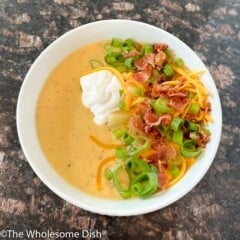 White bowl full of loaded baked potato soup topped with sour cream, green onions, bacon, and cheddar cheese.