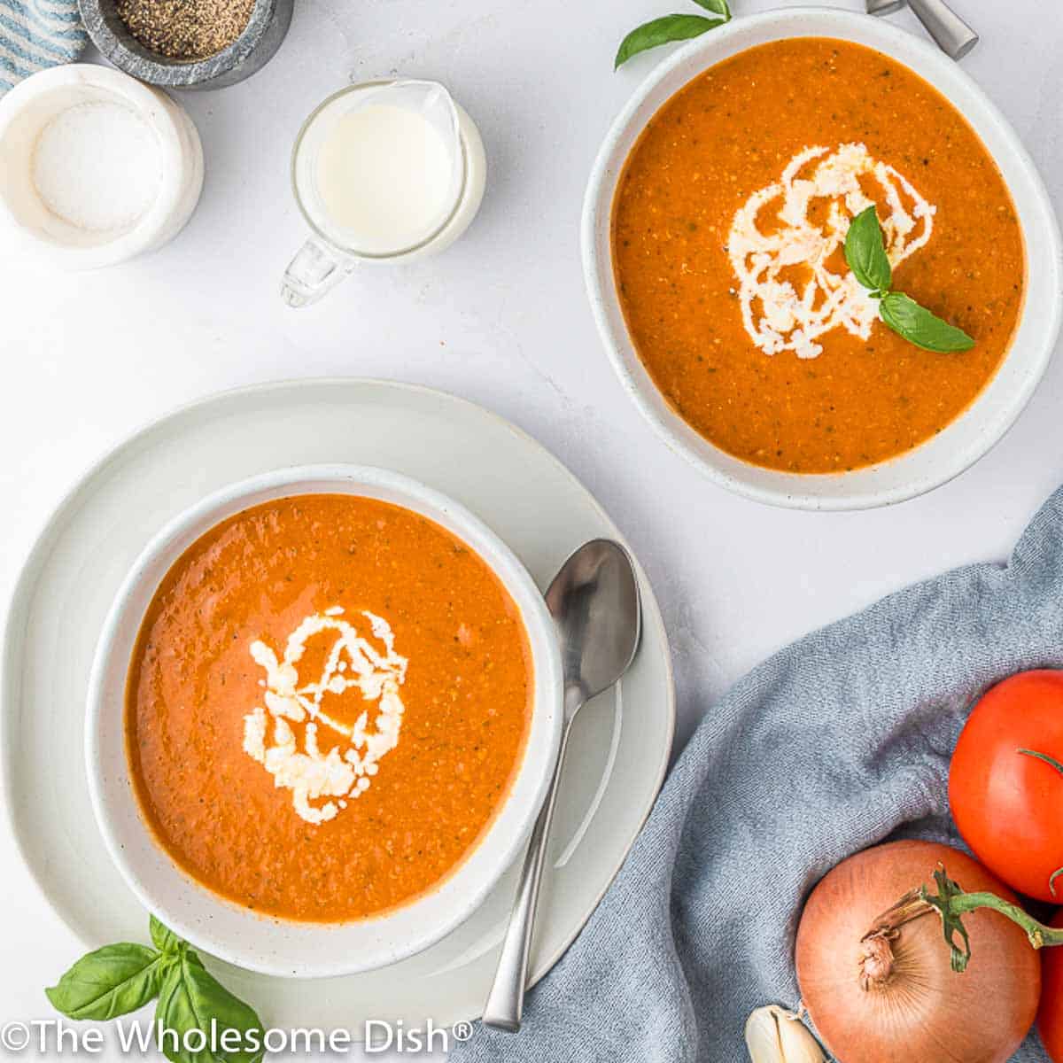 Two white bowls full of roasted tomato soup topped with cream and basil.