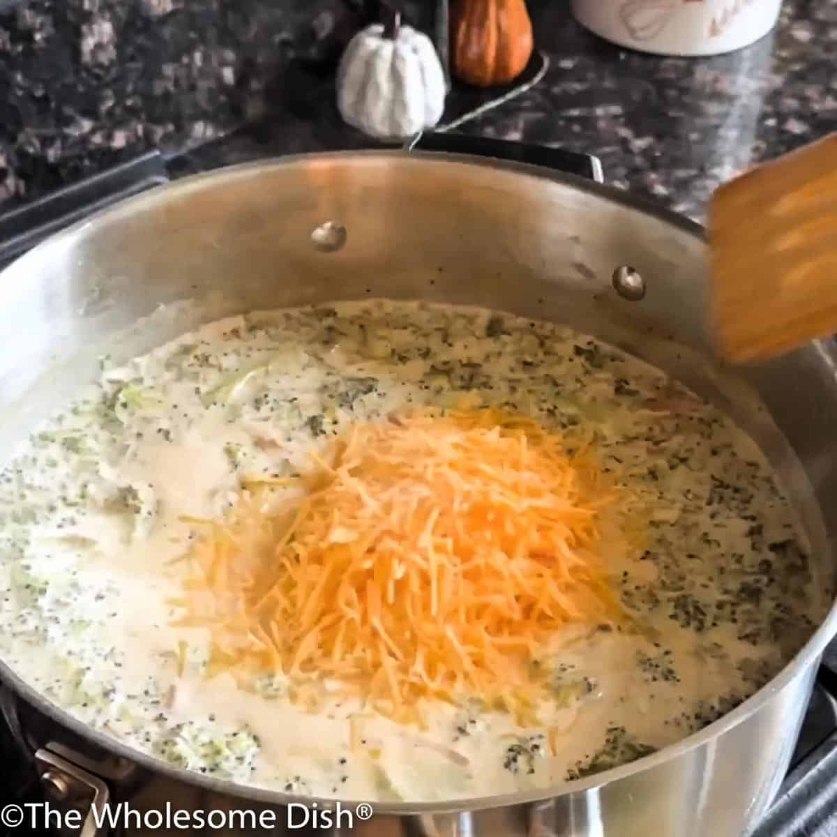 Adding shredded cheddar cheese to the soup pot full of broccoli soup.
