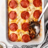 Casserole dish full of Supreme pizza pasta bake with a serving spoon.