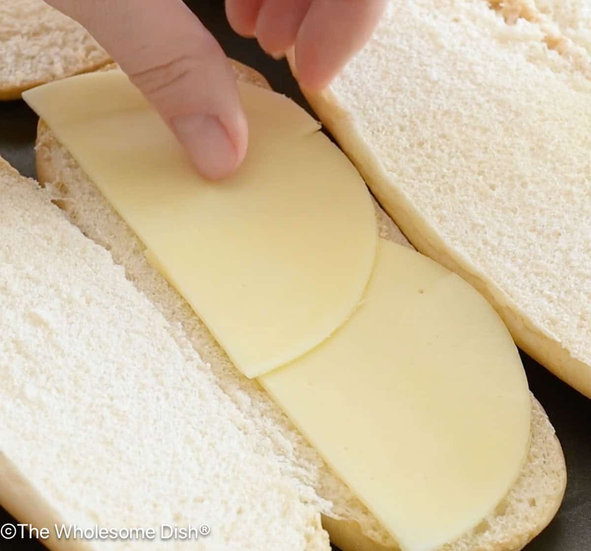 Sliced provolone cheese being added to open sub rolls on a baking sheet.