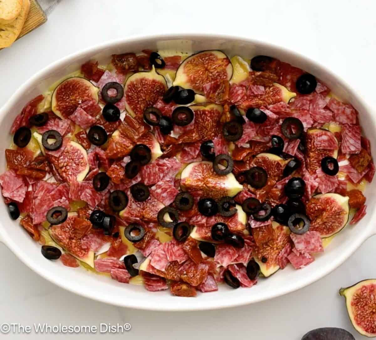 Chopped fig, salami, sun dried tomatoes, and olives over brie and Boursin in a casserole dish.